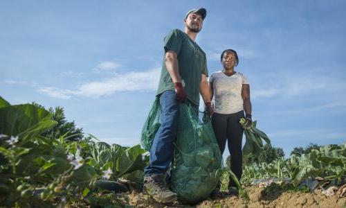 Two farmers stand side by side, both holding crops